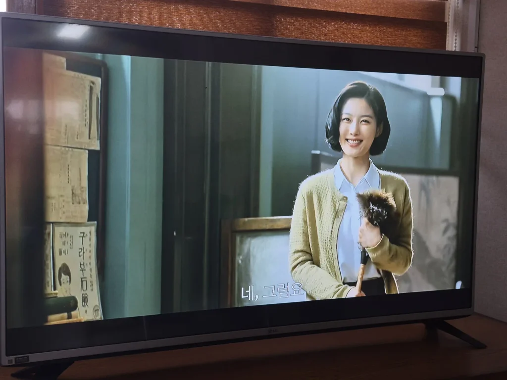 A smiling woman in period-style clothing stands indoors in a scene from 100 Days of Deception on television.