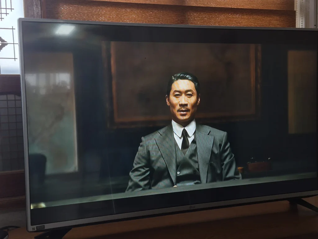A man in a dark three-piece suit sits in a room in a scene from 100 Days of Deception shown on television.